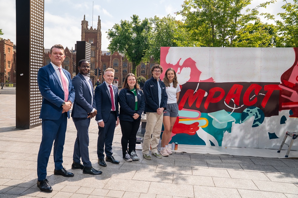 four men and two women smartly dressed and standing on a sunny day beside a large artwork-in-progress showing the word Impact