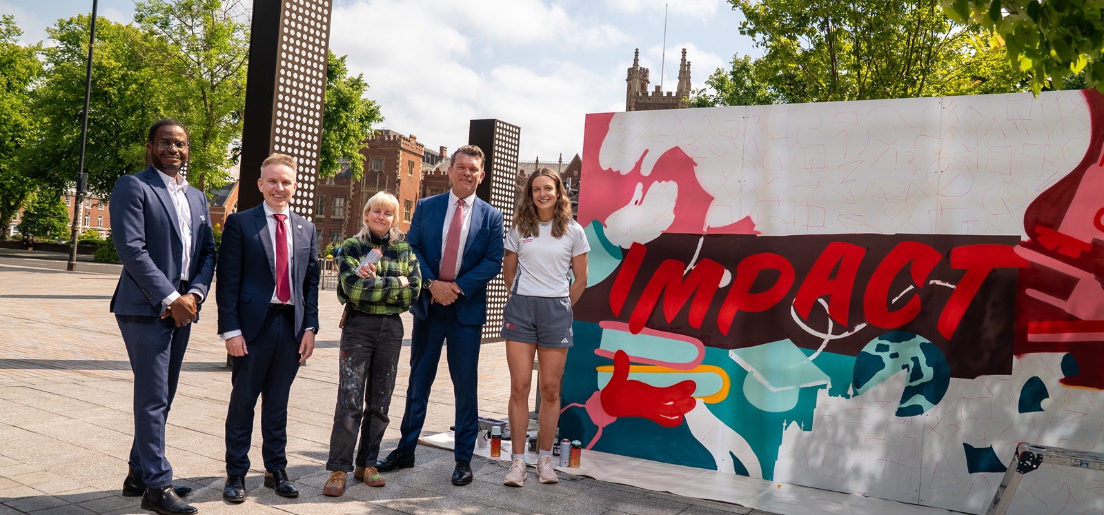 Two women and three men stand posed beside a piece of art which reads the word Impact