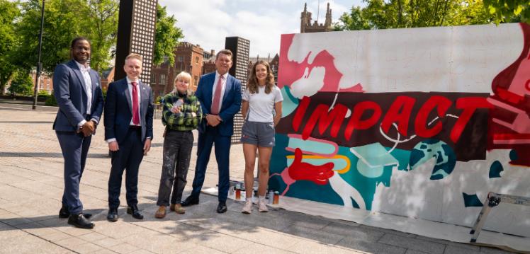 Two women and three men stand posed beside a piece of art which reads the word Impact