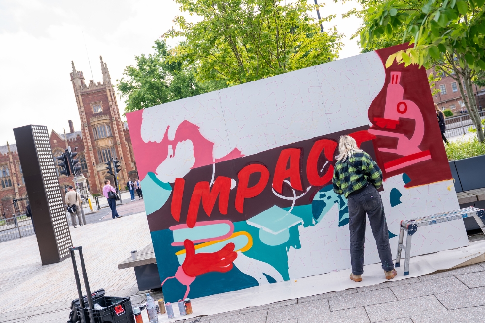 artist creating an artwork outdoors using spraypaint, with old university building and trees in the background