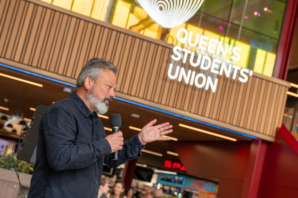 bearded man speaking into a mic at an event with Queen's Students' Union branding shown in the background