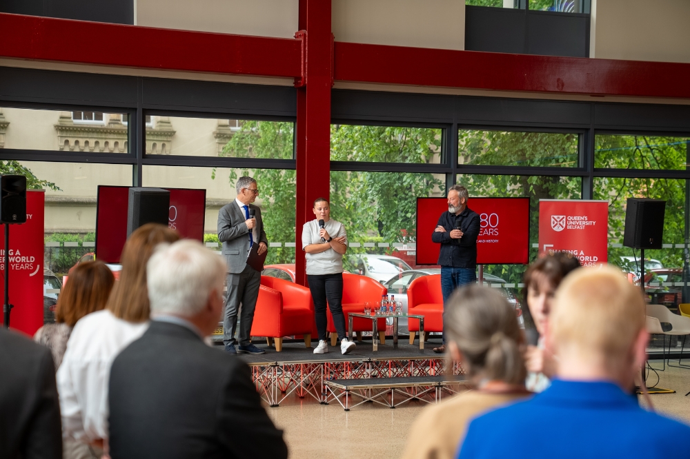 people in foreground gathered and listening to an interview between three people being conducted on a podium in the background