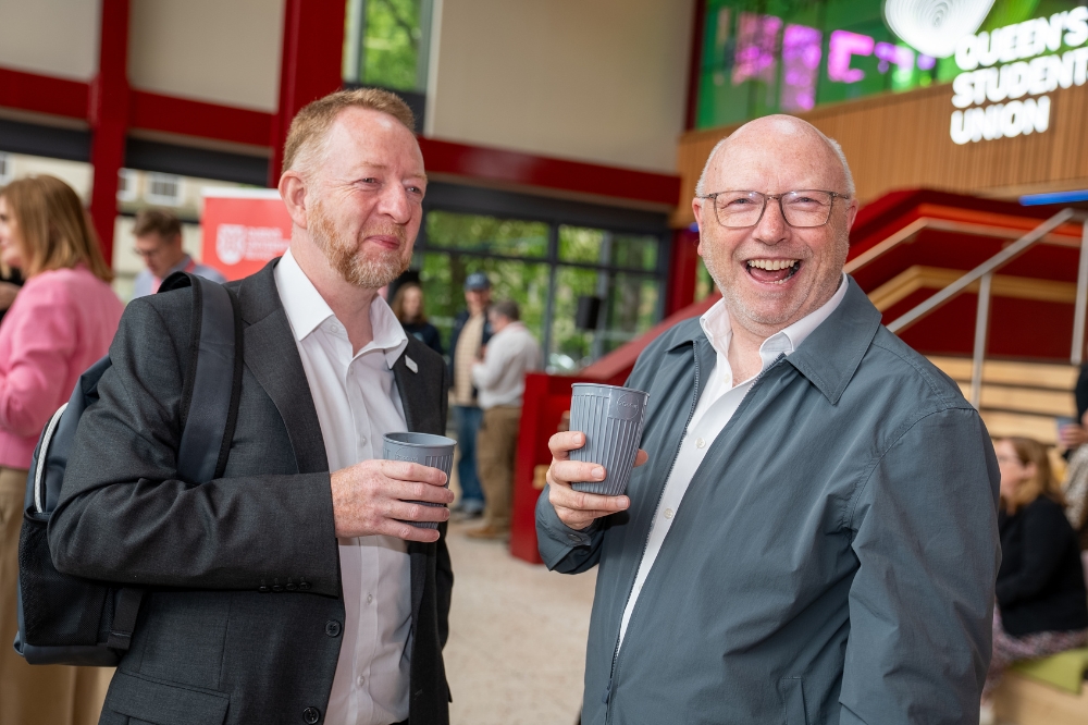 two men holding reusable coffee cups chatting, one smiling to camera