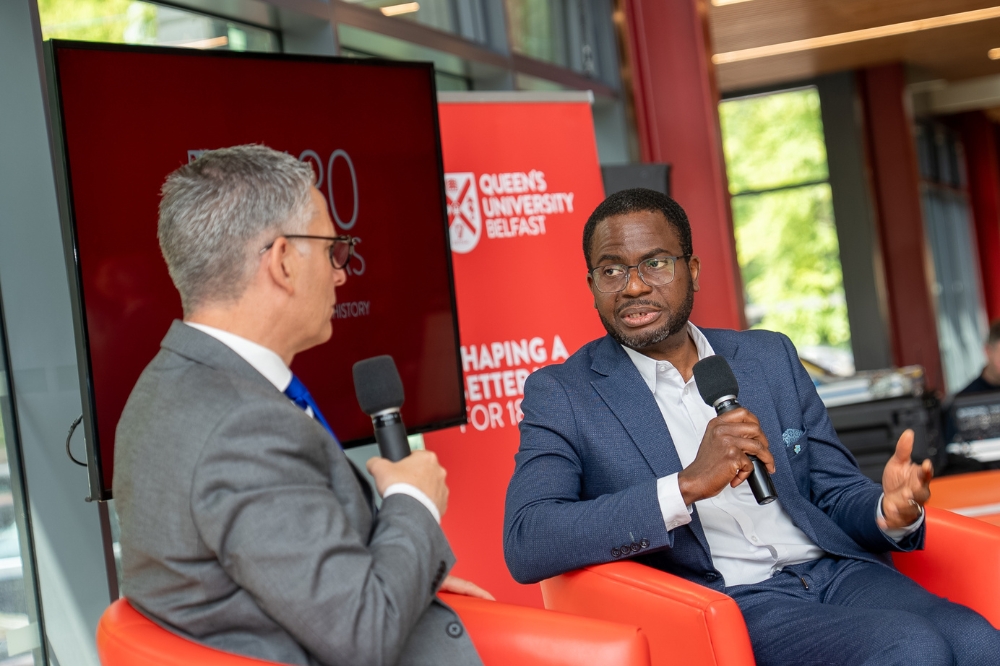 seated, smartly dressed black gentleman being interviewed by another man, with Queen's University Belfast branding shown on pop-up stands in the background