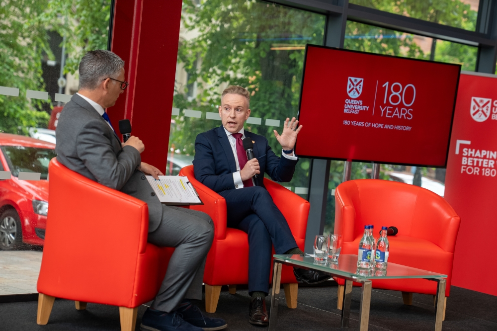 seated, smartly dressed man being interviewed by another man with Queen's University Belfast branding shown on visuals in the background