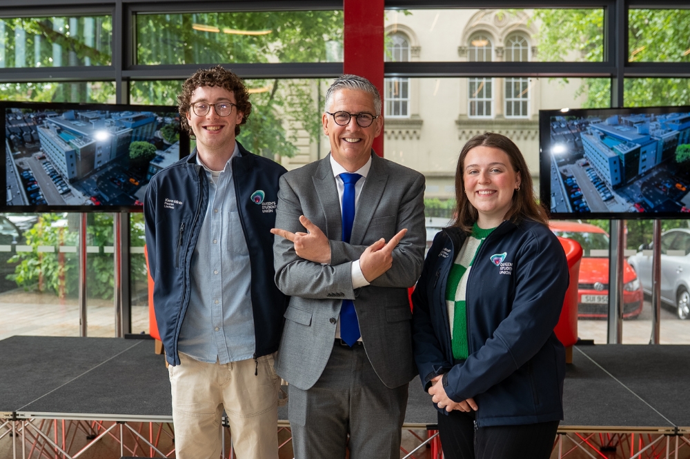 man with glasses standing between and pointing cheerily at a young man and woman on either side of him