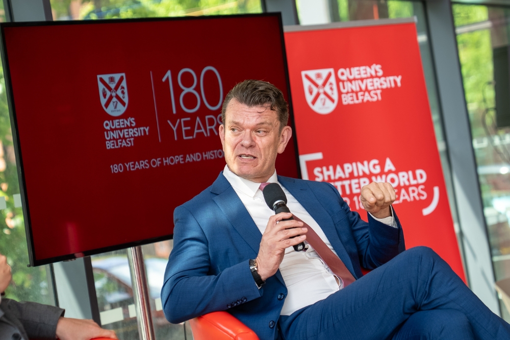seated, well-dressed man speaking into a microphone with Queen's University Belfast branding on pull-up stands in the background
