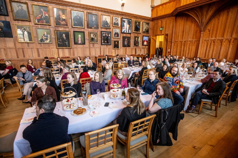 large group of seated people at large tables and listening to an address in an old, wood-paneled indoor space with portraits hanging on the wall in the background