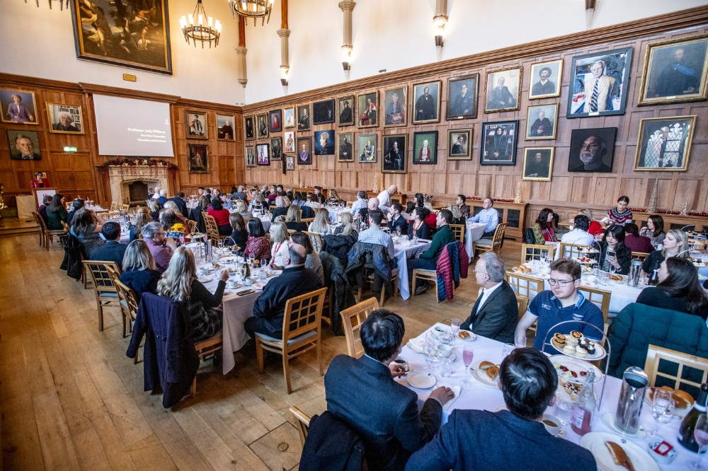 large group of people seated in a large, old, indoor event space with portraits hanging on the far wall