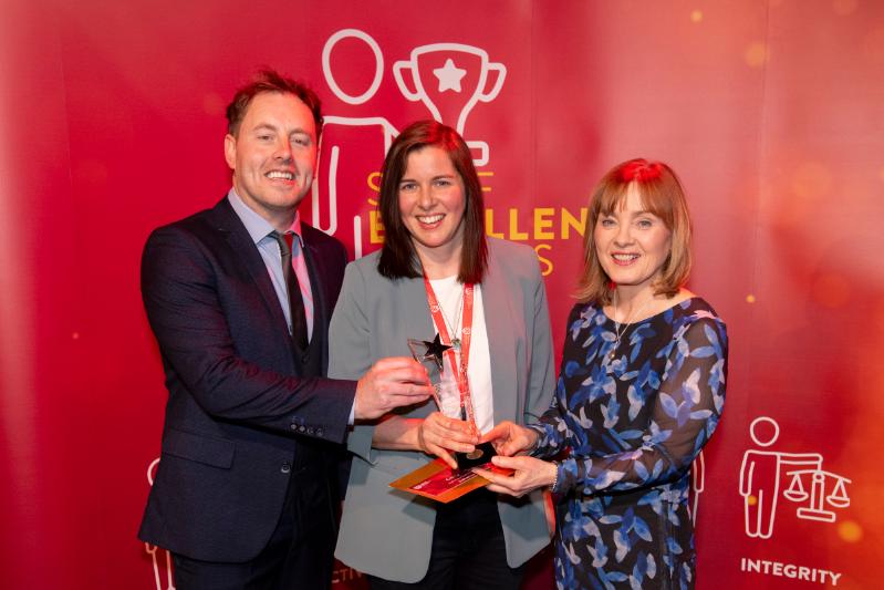 A man and three women standing in front of a large red Staff Excellence Awards banner.  Lady in the middle is hold a glass trophy award