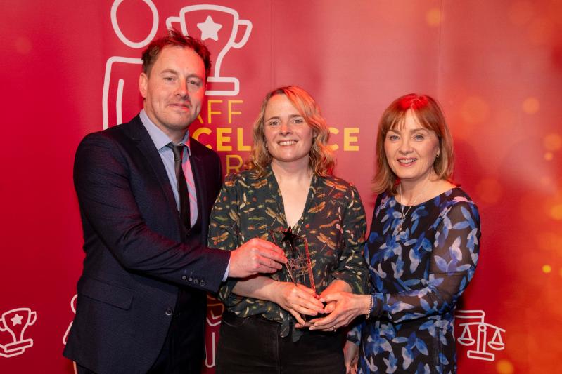 A man and two women standing in front of a large red Staff Excellence Awards banner.  Lady in the middle is hold a glass trophy award