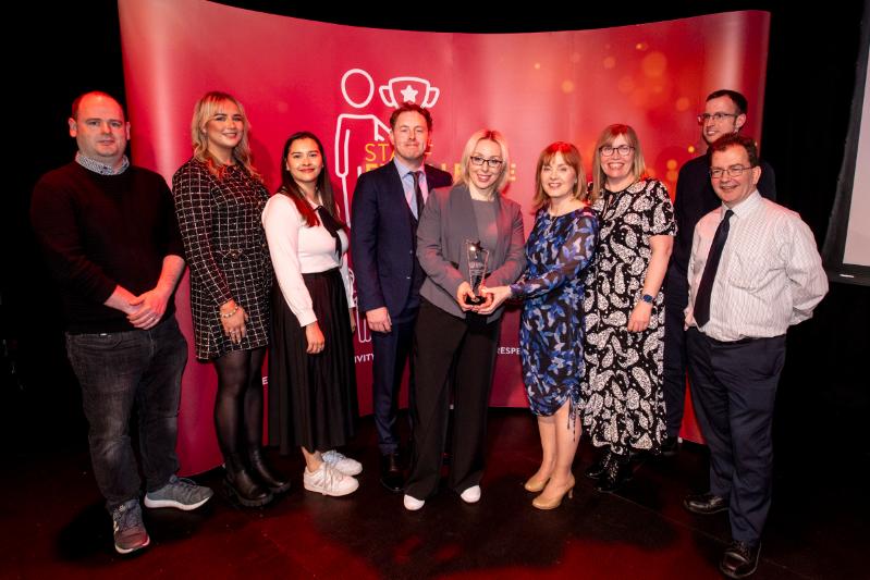 nine adults standing in front of a red Staff Excellence Awards Banner, with lady in the middle recieving a glass trophy award