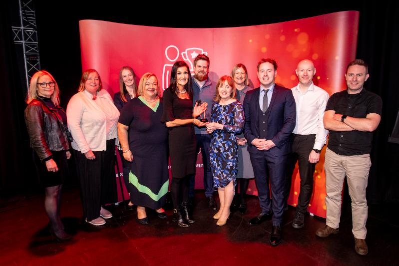 Eleven adults standing in front of a red Staff Excellence Awards banner with fifth lady in the middle receiving a glass trophoy award
