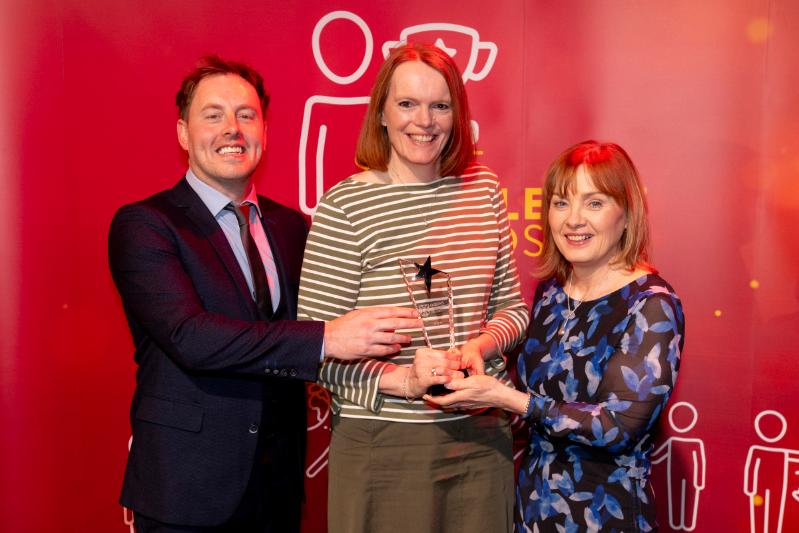 On man and and two ladies stading in front of a red Staff Excellence Awards banner, with lady in the centre receiving a glass trophy award