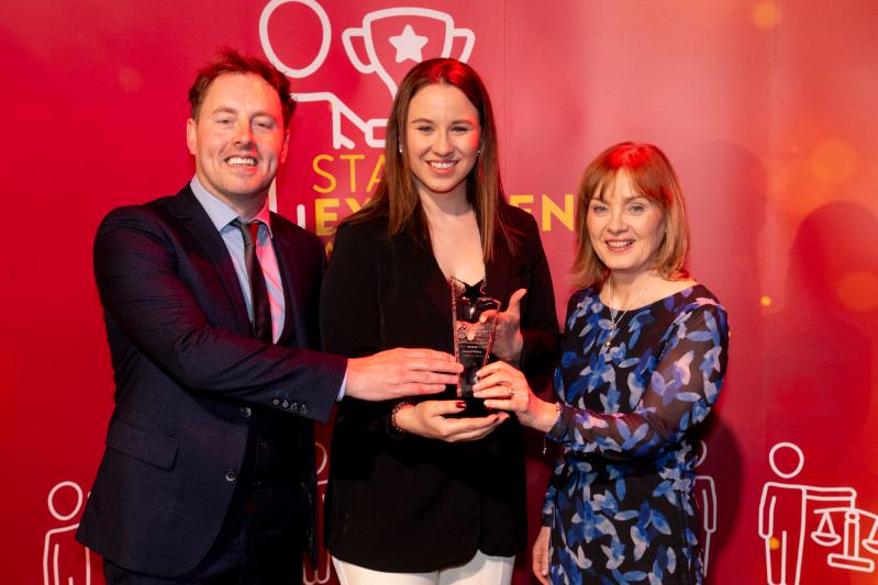 One male and two female standing in front a red Staff Excellence Awards banner, the lady in the centre receiving a glass trophy award