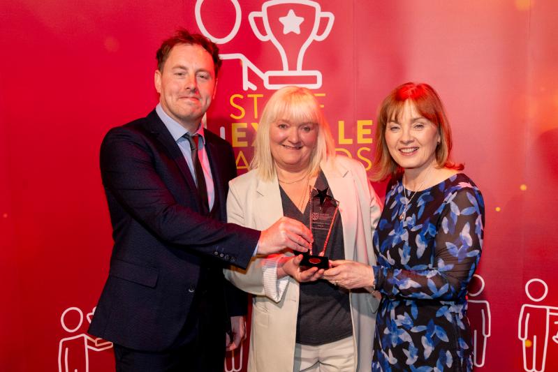 On man and and two ladies standing in front of a red Staff Excellence Awards banner, with lady in the centre receiving a glass trophy award