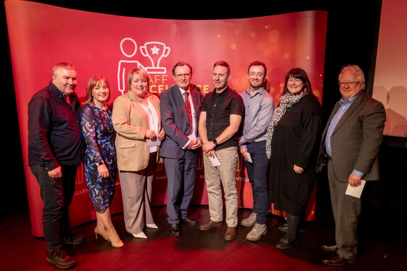 Eight QUB members of staff, including the VC standing in front of a red Staff Excellence Awards BAnner