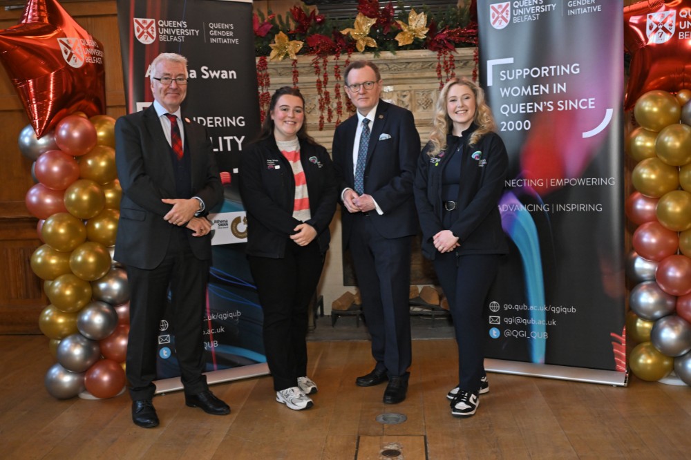 two mature men wearing glasses and suits and two young women posing for a photo in an old wood-paneled event hall with large decorated fireplace and pop-up stands in the background