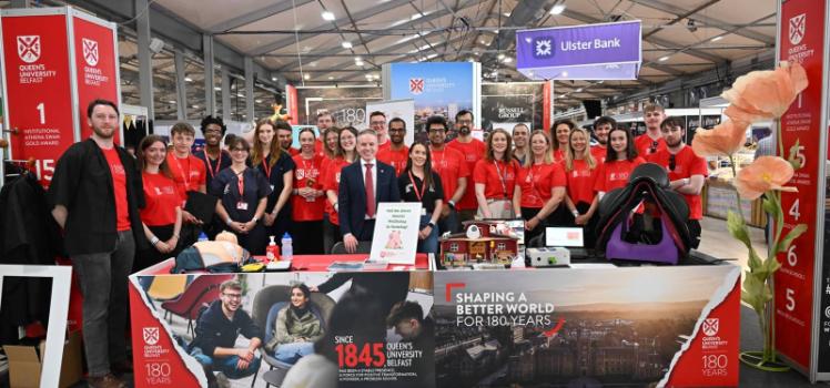 large group of smiling people at a Queen's University Belfast-branded indoor exhibition stand