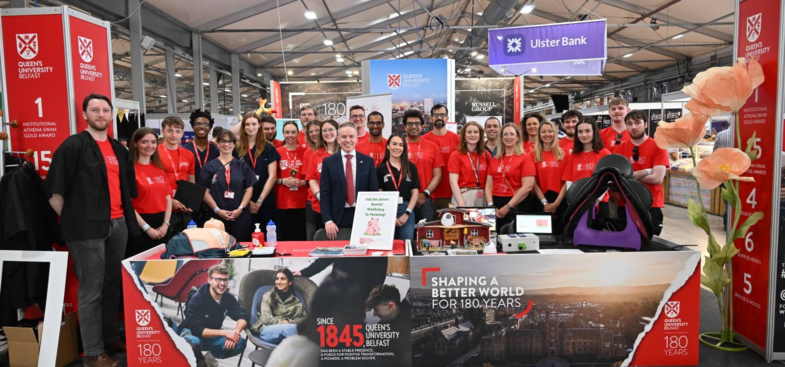 large group of smiling people at a Queen's University Belfast-branded indoor exhibition stand