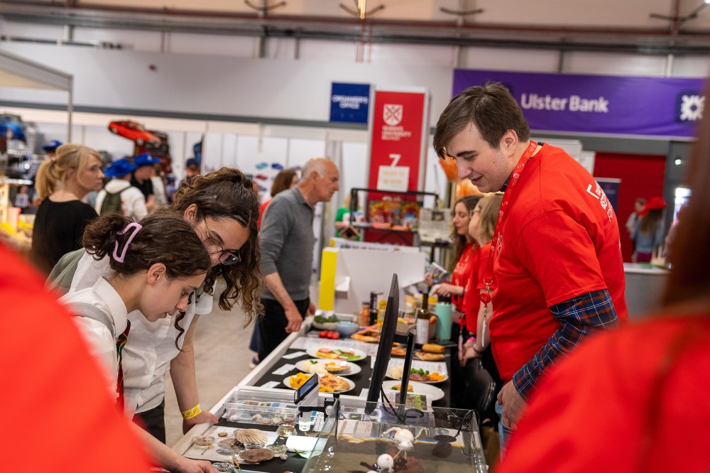 two girls looking at exhibits and a man in red t-shirt on hand to help at an exhibition stand