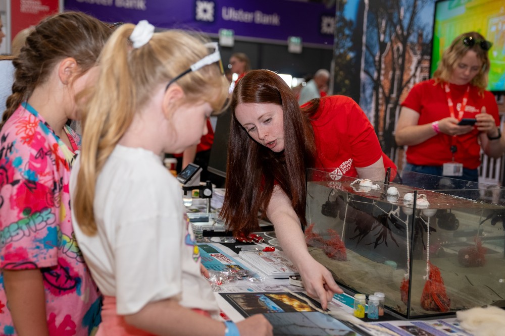 young woman in red t-shirt standing behind a marine life tank at an exhibition stand, talking to and showing information to two children