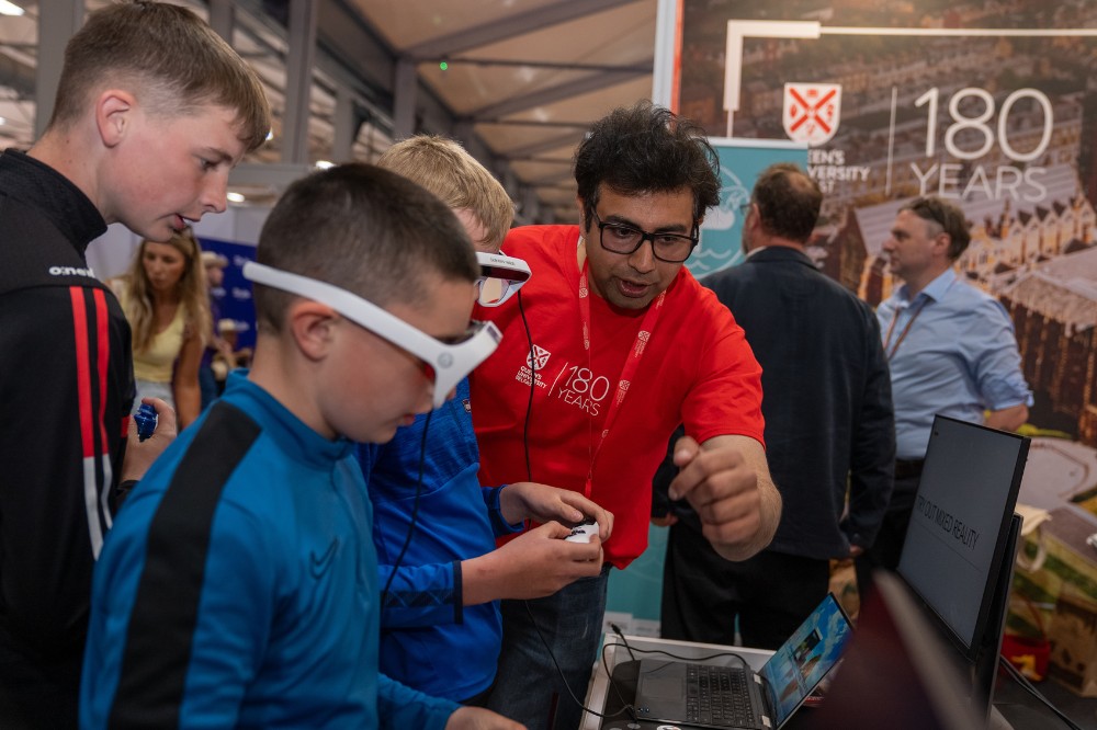 man in red t-shirt demonstrating a VR headset to three boys at an exhibition stand