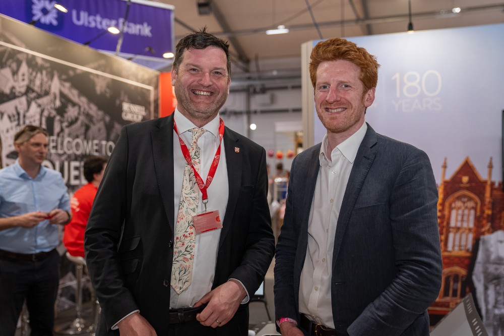 two smartly dressed, smiling men - one wearing a red lanyard - at an exhibition stand