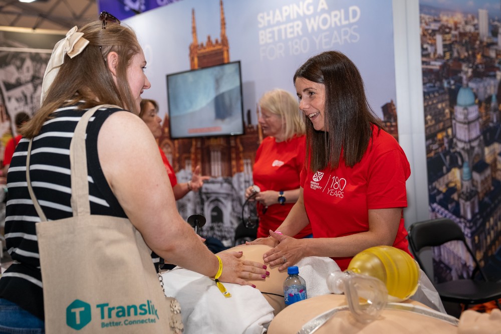 woman in red t-shirt demonstrating simulation equipment to a female passer-by at an exhibition stand