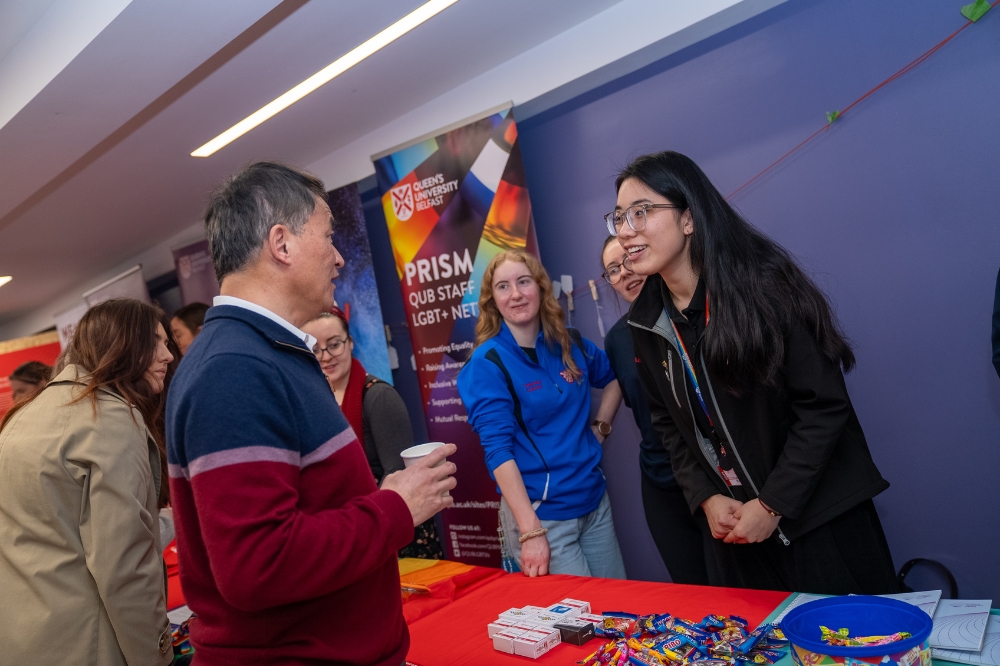 People talk and smile at a PRISM staff network stall during an indoor campus safety event, with information materials on the table.