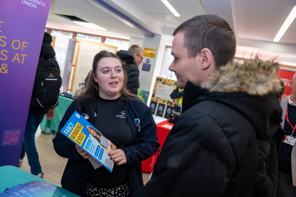 Student Union President Amy Smith speaks with an attendee at an indoor campus event while holding an information leaflet.