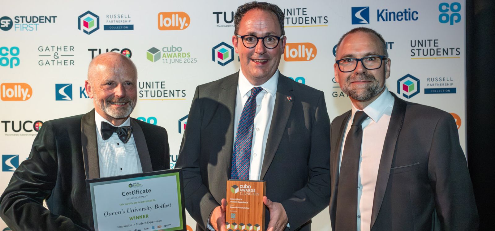 Three suited male Queen's staff standing in front of an advertising banner holding the CUBO award trophy and certificate for Innovation in Student Experience