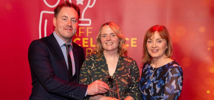 Three people standing together at an awards event, smiling and holding a trophy, with a red background displaying the words ‘Staff Excellence Awards.
