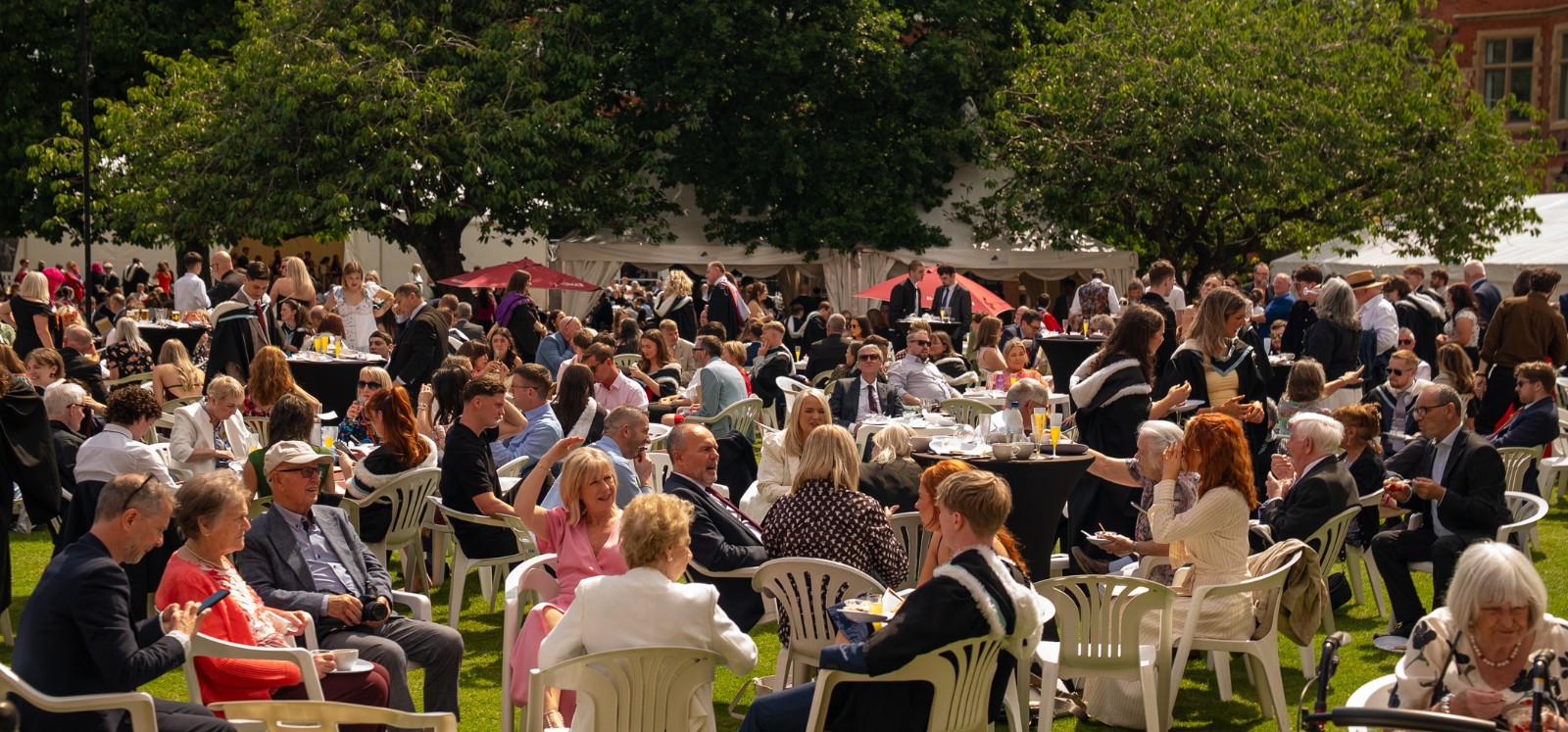groups of people in formal wear sitting on a lawn enjoying refreshments, with trees in the background
