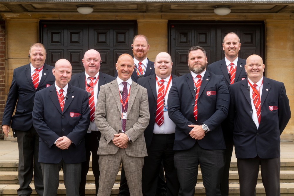 group of eight men in matching navy-blue suits, and another wearing grey, standing with hands folded outside the entrance to a building