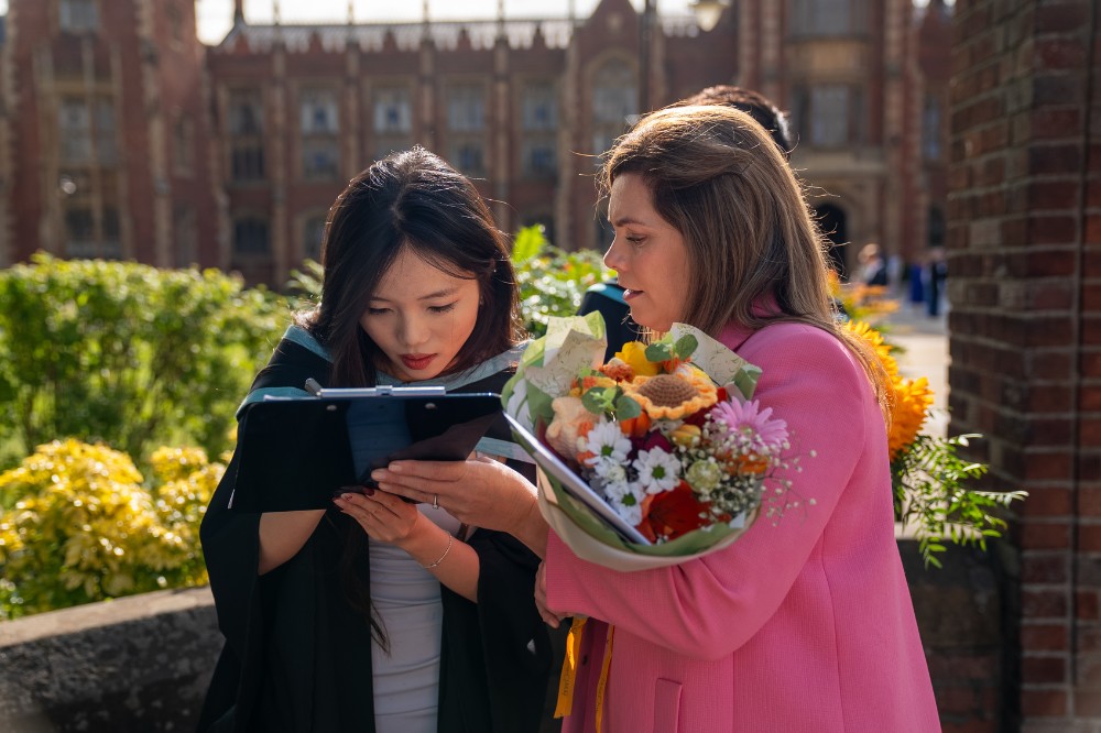 young female graduating student writing on a notepad with another woman to her right in a pink jacket holding a bunch of flowers