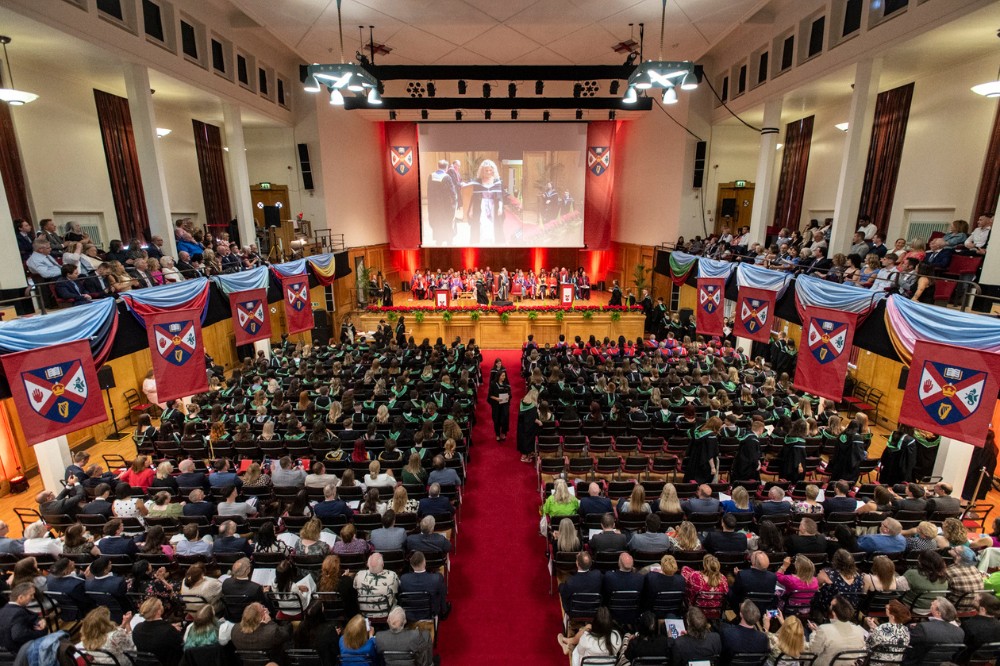 view from upper floor looking towards the stage of a large hall filled with graduating students, robed academic staff, friends and families