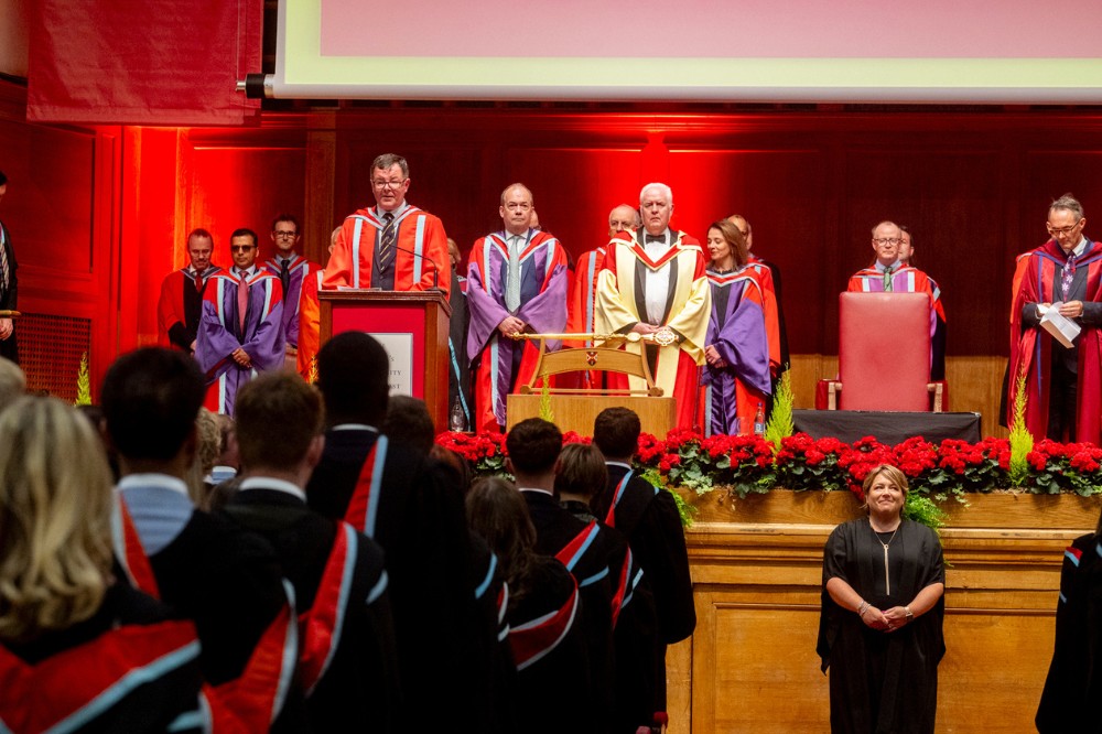 graduands and academics in graduation robes standing for a citation in a large hall