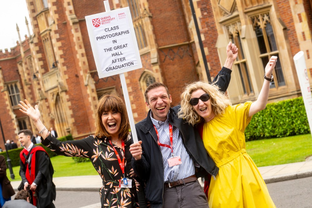 two smiling, celebrating women flanking a man holding a placard that reads 'Class photographs in the Great Hall after graduation'