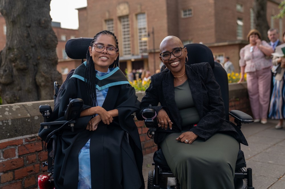 two smiling women in wheelchairs, one wearing a graduation robe, with an old building in the background