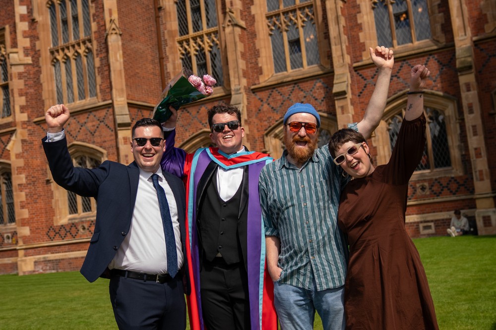 three men and a lady raising hands in celebration on a sunny day outside a redbrick building