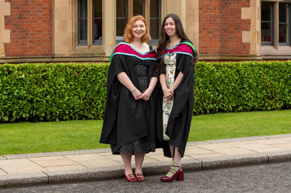 two young women in graduation robes standing on a pavement and smiling to camera