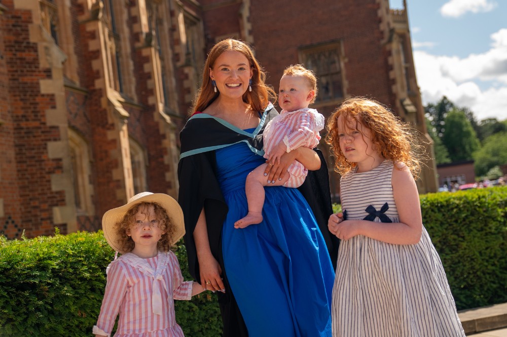 young woman in graduation robe posing with three children outside a redbrick building in the sunshine