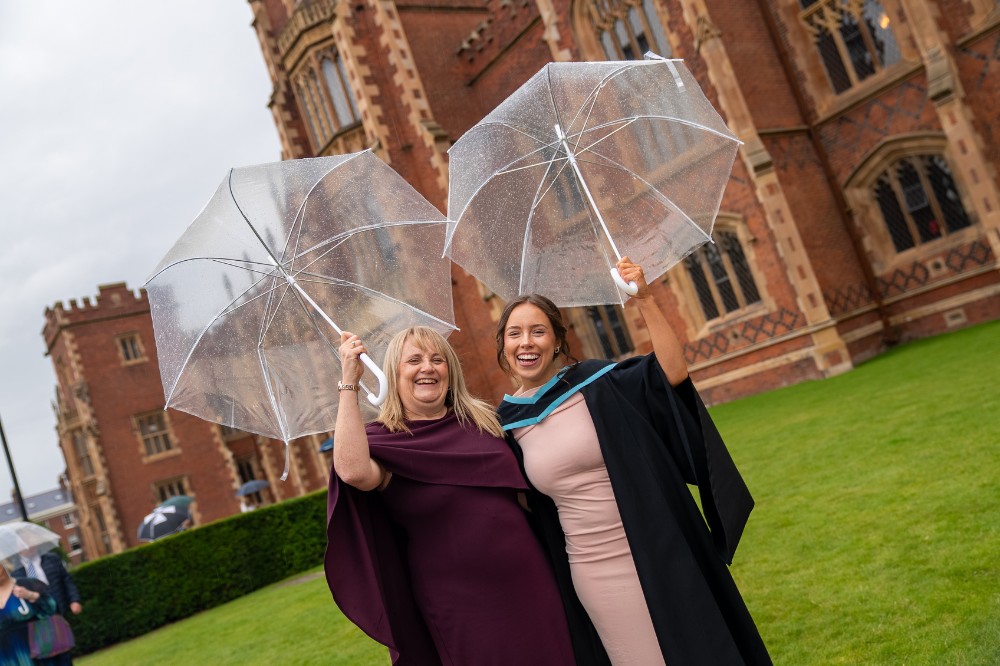 two smiling women holding umbrellas on a lawn outside a redbrick building - the woman on the right is wearing a graduation robe
