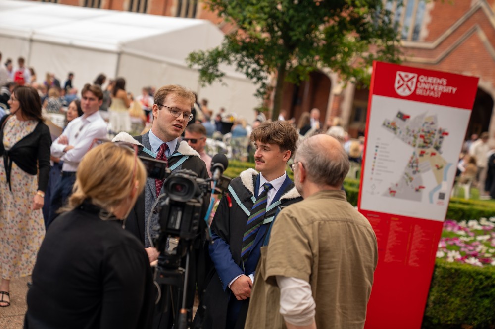 two male graduating students talking to man and woman operating a film camera with a graduation party taking place in the background