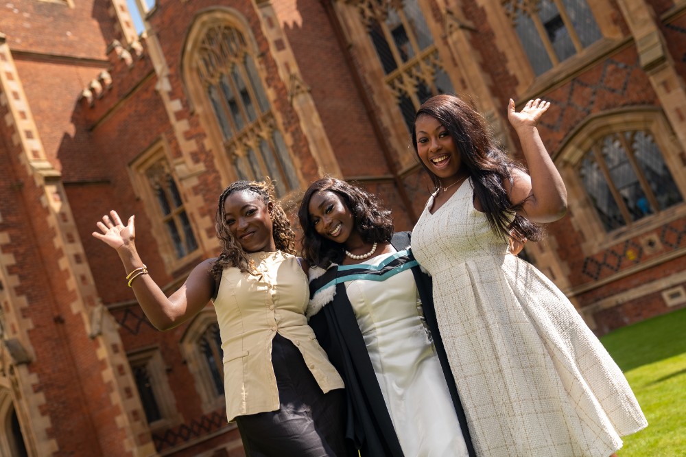 three women of African origin smiling and waving to camera outside a redbrick building in the sunshine
