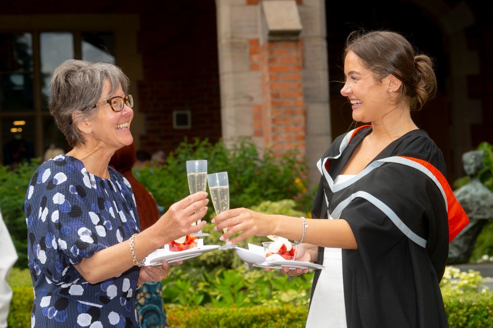 a mother and daughter wearing graduation robe clinking glasses at a garden party
