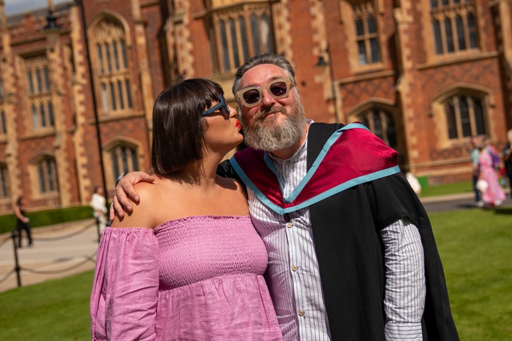 man in thick-rimmed sunglasses and graduation robe receiving a kiss from his partner in the sunshine with a redbrick building in the background