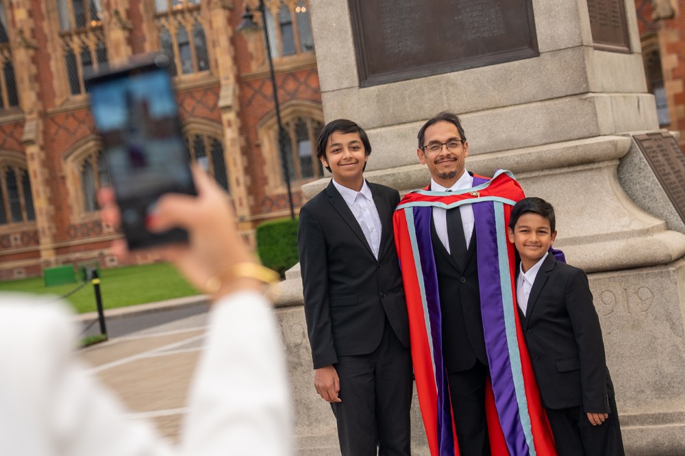 man in graduation robe posing with two children in front of a monument and posing for a photo someone is taking on a phone in the foreground