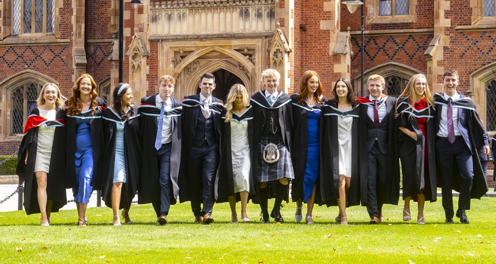 group of graduating young men and women lined up side by side and walking towards the camera for a photo op on a lawn in the sunshine with a redbrick building in the background